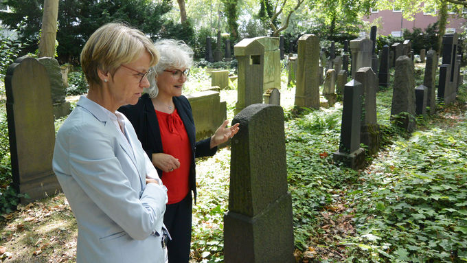 zwei weibliche Personen stehen auf einem Friedhof mit mehreren Grabsteinen