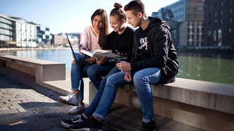 Jugendliche sitzen auf einer Mauer an einem Hafen und schauen in ein Laptop