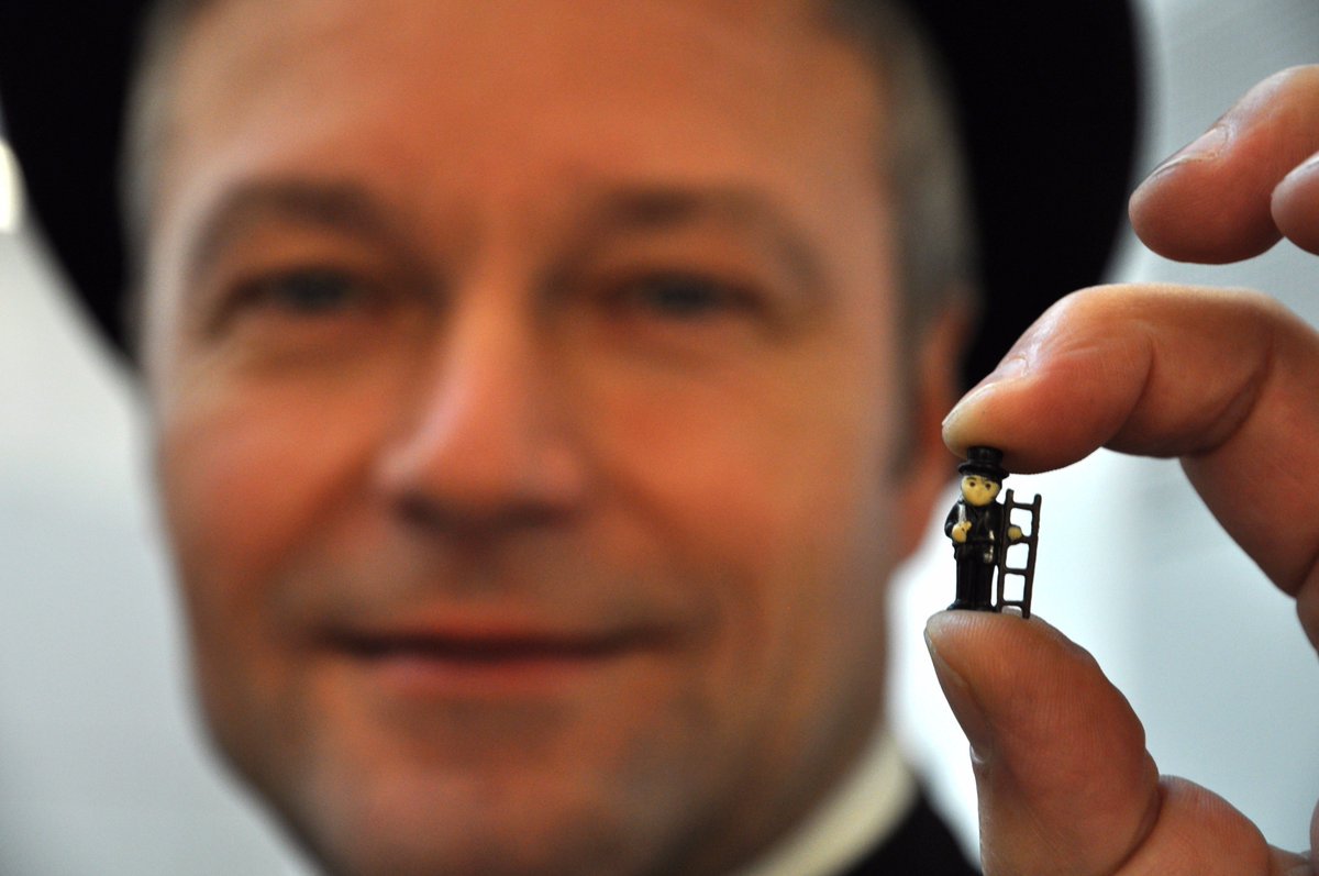 Gesicht eines Schornsteinfegers im Hintergrund unscharf, im Vordergrund hält er einen kleinen Plastikschornsteinfeger in die Kamera