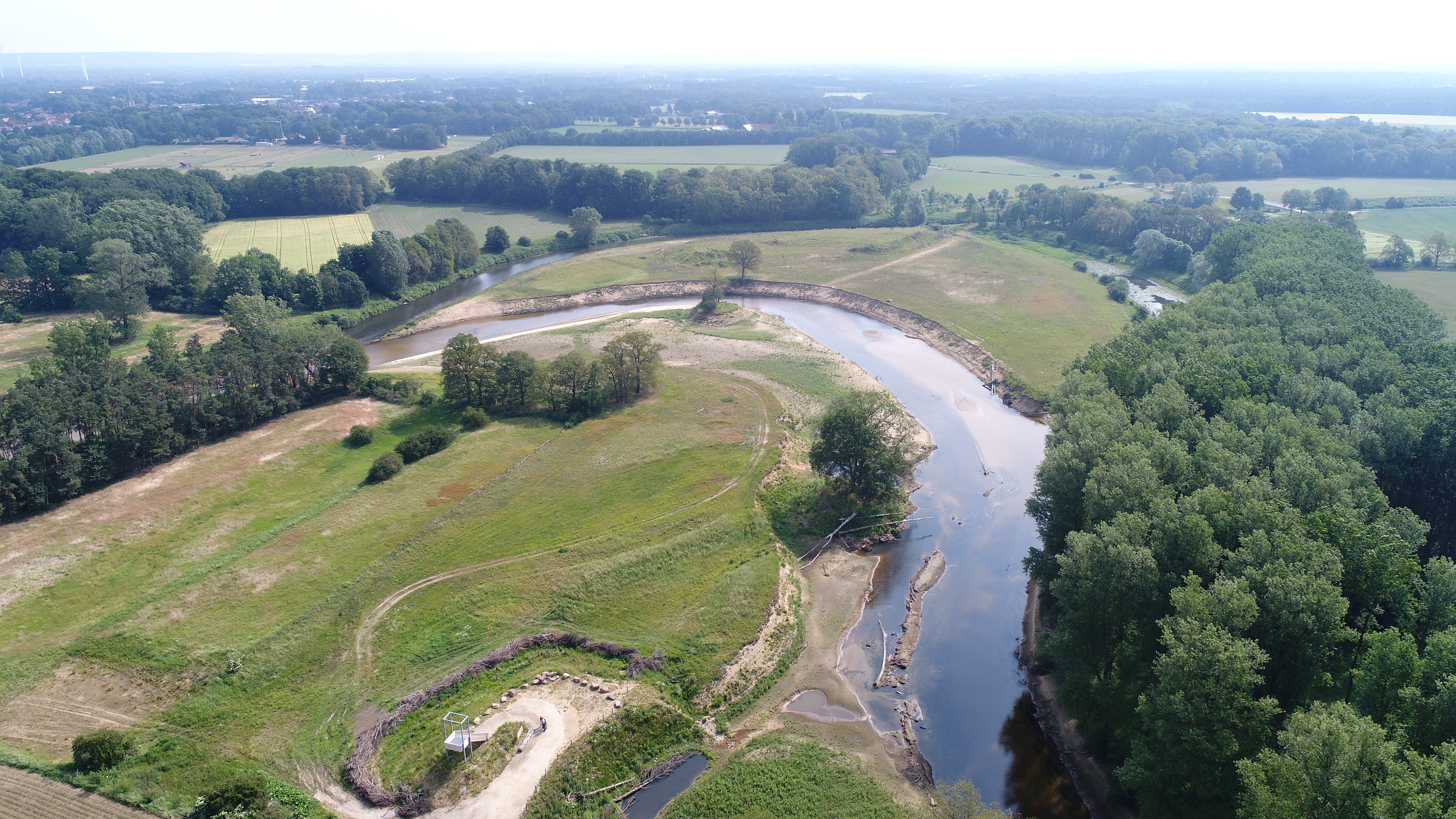 Zu sehen ist eine Drohnenaufnahme des nördlichen Abschnitts des wieder an die Ems angebundenen Altarms Hembergen. Im linken Bildbereich mündet der Altarm wieder in den bestehenden Emsverlauf. Im hinteren Bildbereich ist der ausgebaute Emsverlauf zu sehen, der bestehen bleibt und sich als Altgewässer entwickeln darf. Im Vordergrund befindet sich ein Aussichtsturm.