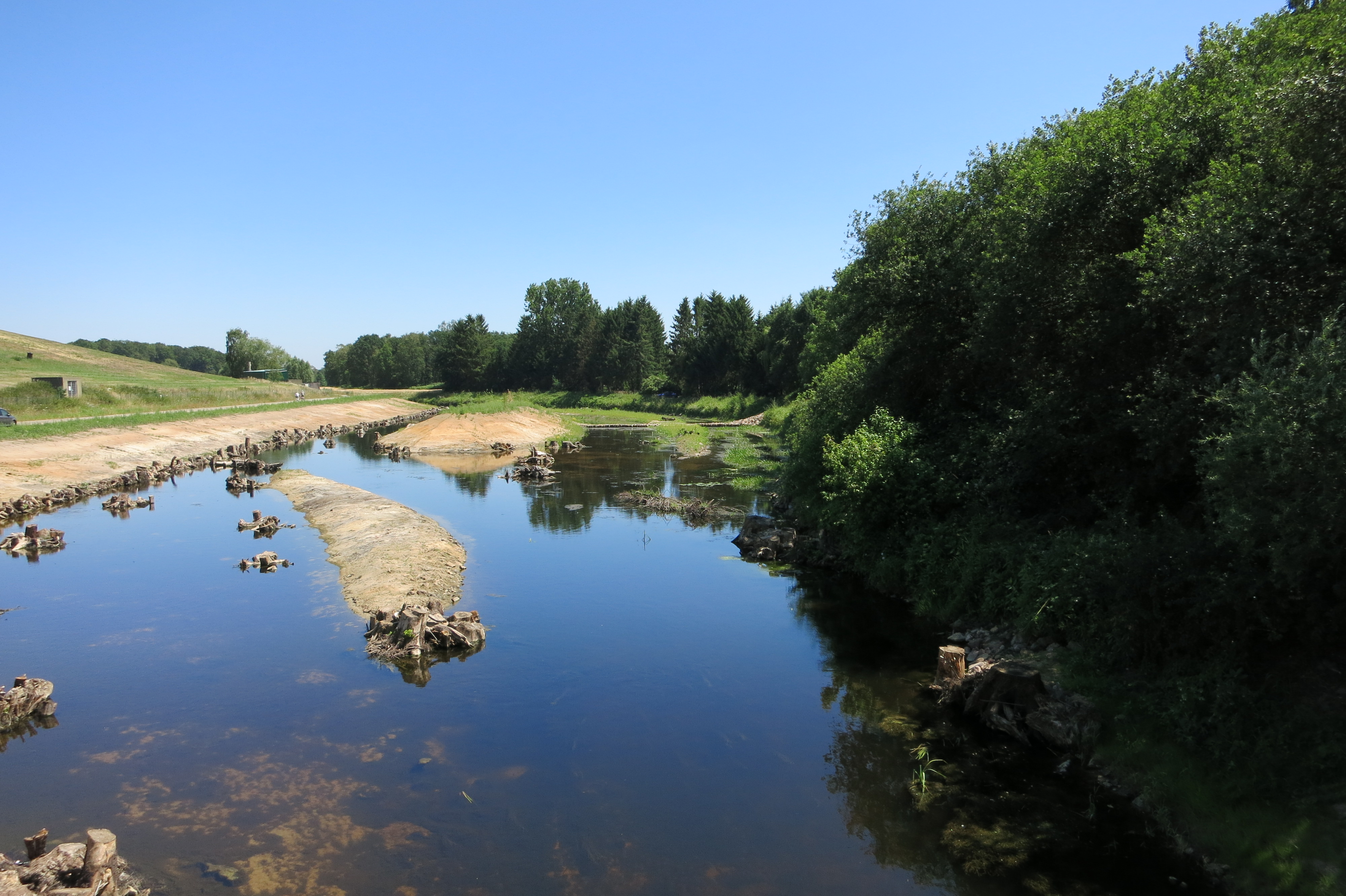 Zu sehen ist ein Abschnitt der Bocholter Aa in Hoxfeld, in dem strukturverbessernde Maßnahmen umgesetzt wurden. Mehrere Wurzelstubben wurden eingebaut und Inseln angelegt, um die Strukturvielfalt zu erhöhen. Links angrenzend an die Bocholter Aa ist die Deponie Hoxfeld zu erkennen, am rechten Ufer ist ein Gehölzsaum entlang der Aa zu sehen. 