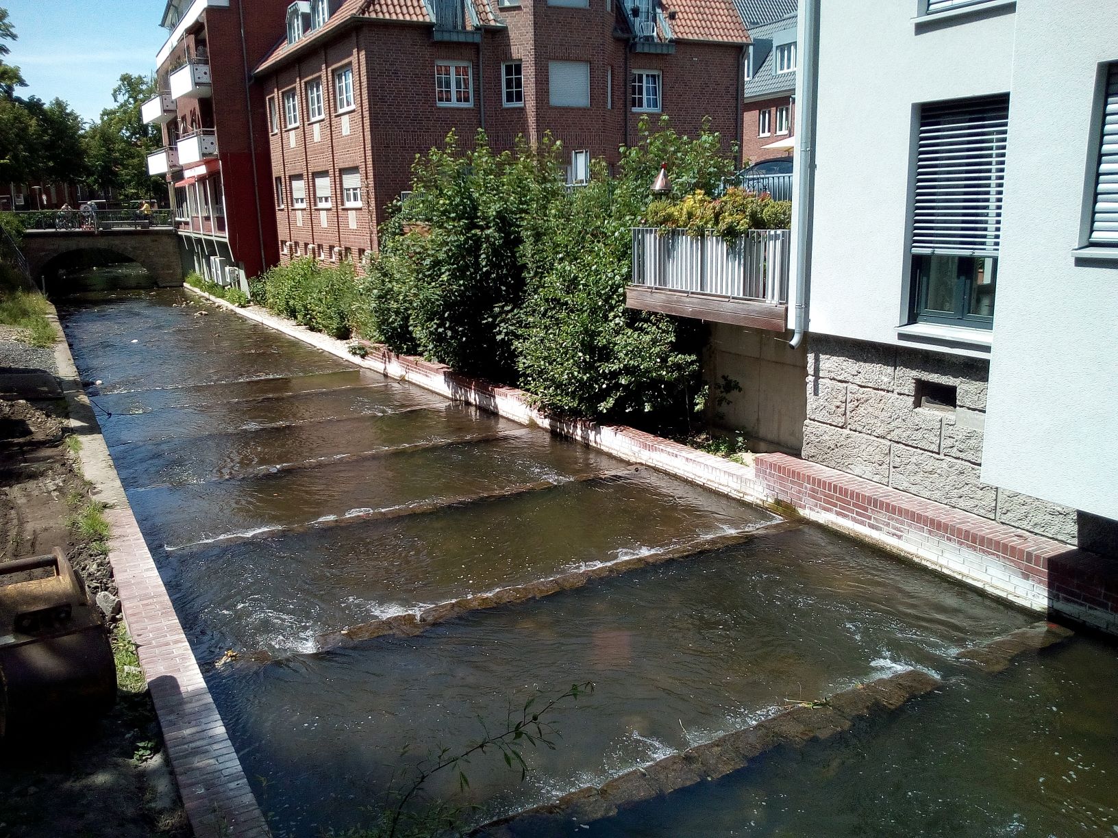 Zu sehen ist die etwa 8 m breite Berkel in der Innenstadt von Coesfeld. Beide Ufer werden von niedrigen Mauern gebildet. Auf der in Fließrichtung rechten Seite grenzt Wohnbebauung mit abschnittsweise vorgelagerten niedrigen Gebüschen an die Berkel an. Im Gewässerbett ist die Sohlgleite mit ihren einzelnen Steinriegeln und Öffnungen zu erkennen, die das ehemalige Wehr ersetzen und den Organismen die Durchwanderbarkeit der Berkel ermöglichen. 