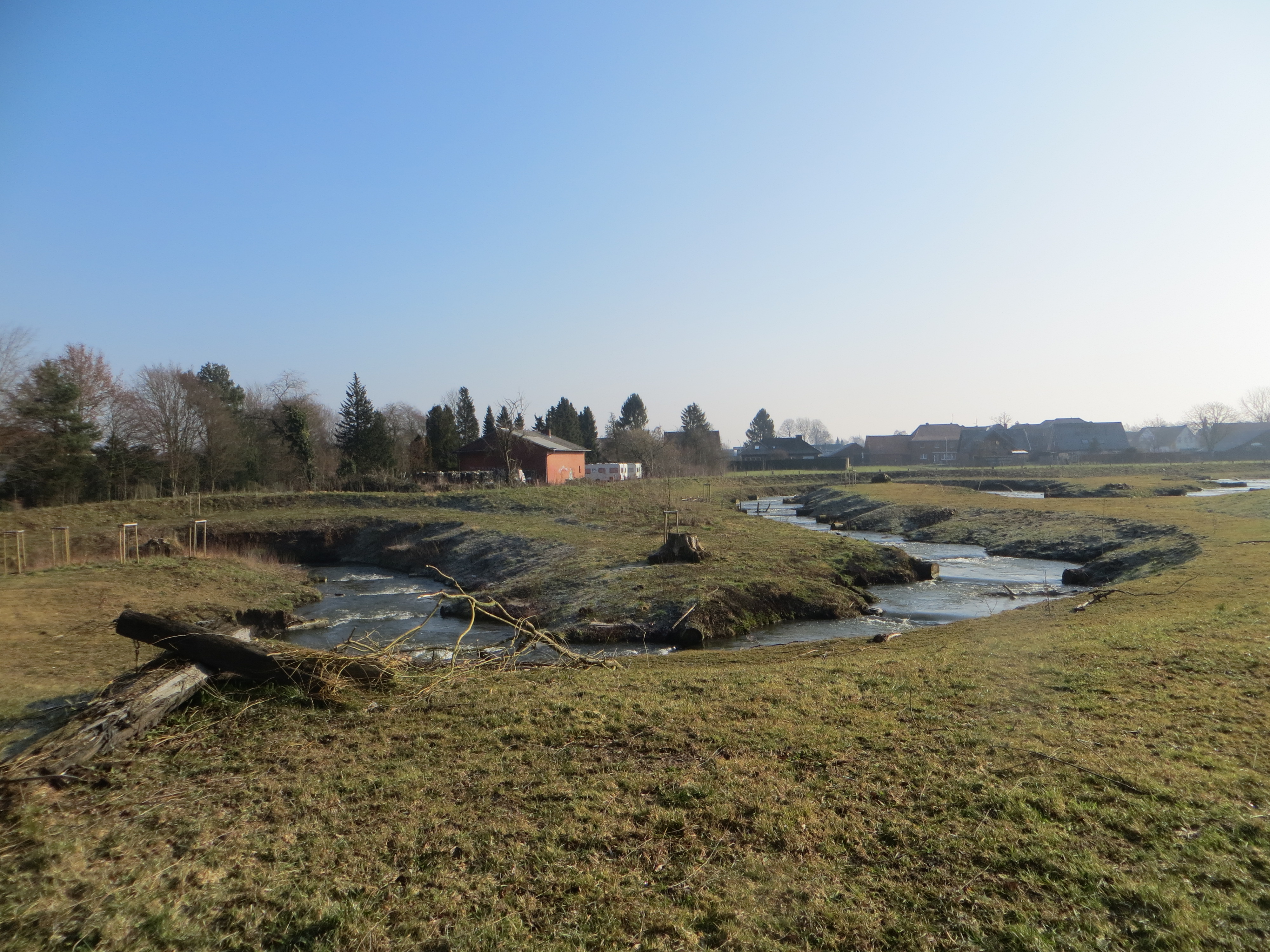 Zu sehen ist ein in Mäandern angelegtes Umgehungsgerinne zur Herstellung der Durchgängigkeit an der Plagemanns Mühle in Metelen in Riegelbauweise. Im Hintergrund ist die Wohnbebauung von Metelen zu sehen. 