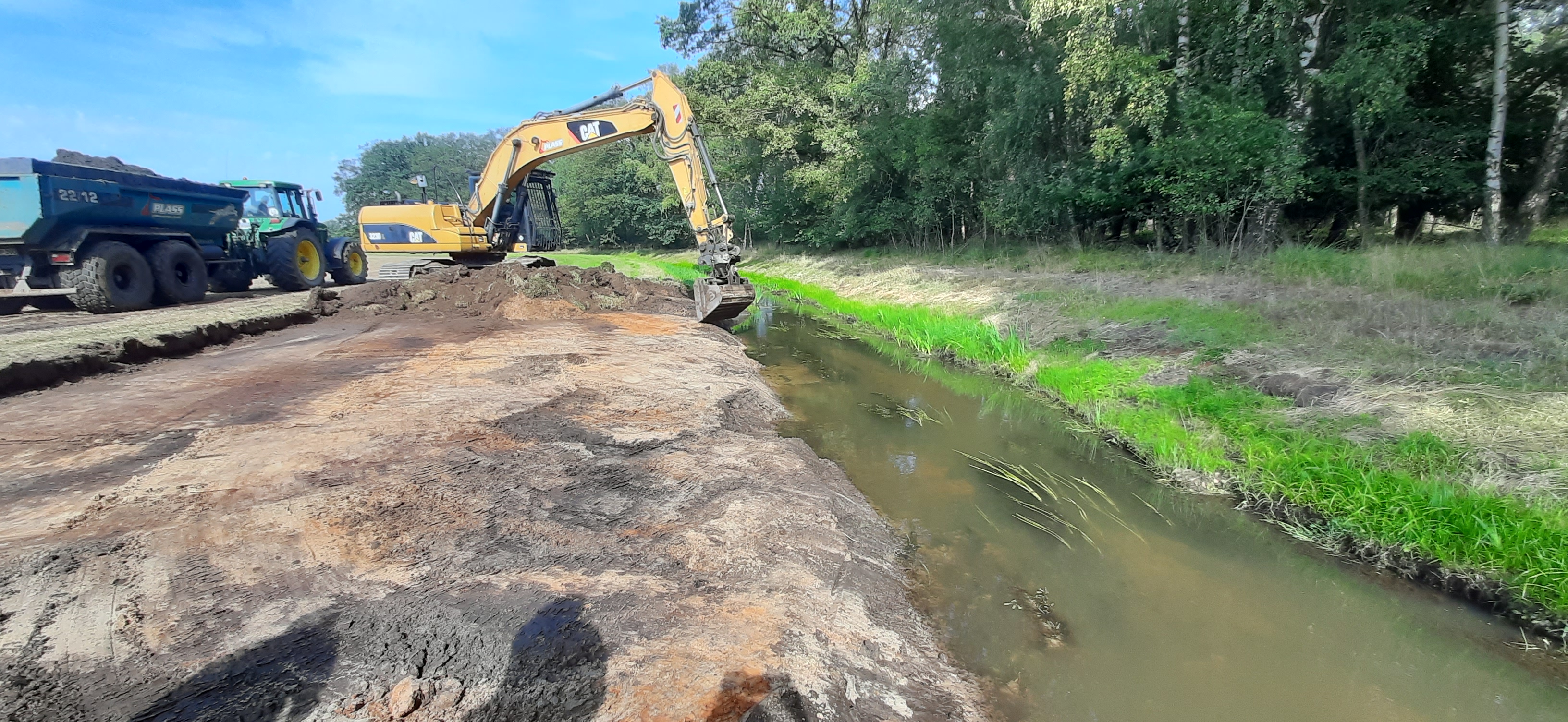 Ein Bagger nimmt Abgrabungen am Ufer eines Baches vor. Hinter dem Bagger befindet sich ein Traktor mit Anhänger, der die Erde abtransportiert. 