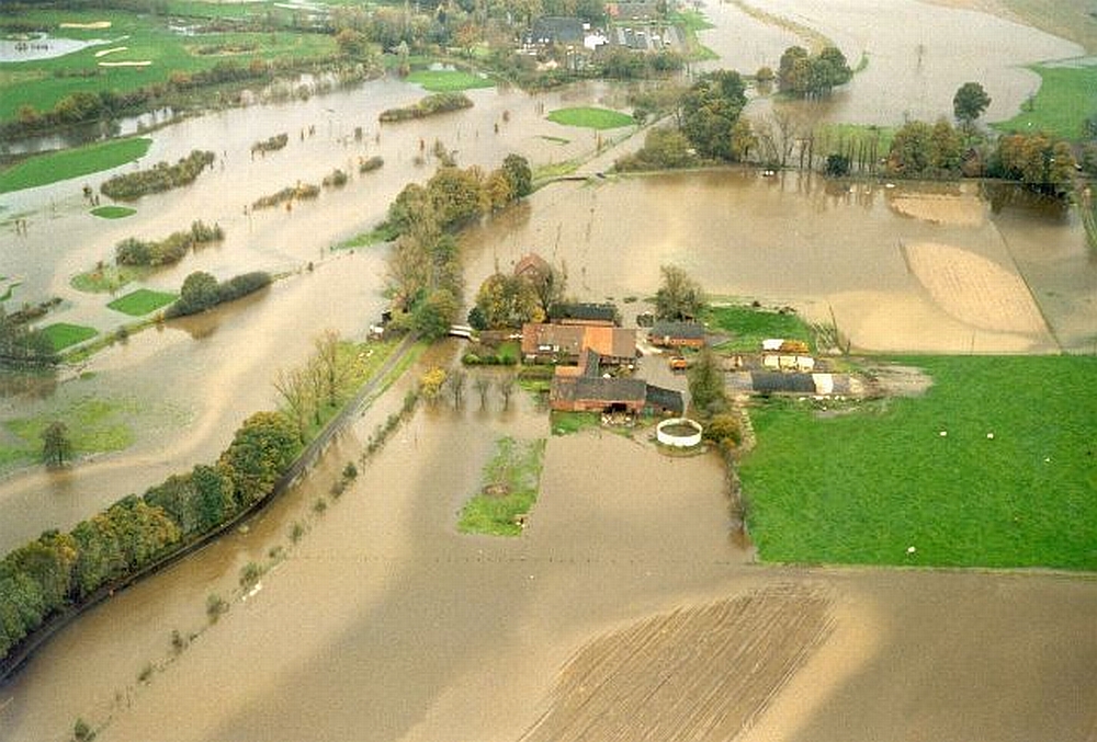 Das Luftbild zeigt einen Fluss, der über die Ufer getreten ist und weite Teile der umliegenden landwirtschaftlich genutzten Flächen überschwemmt. Mittig des Bildes ist eine Gebäudeansammlung zu sehen, bei dem es sich um die Kläranlage Isselburg handelt. Diese ist vollständig von den Wassermassen eingeschlossen.