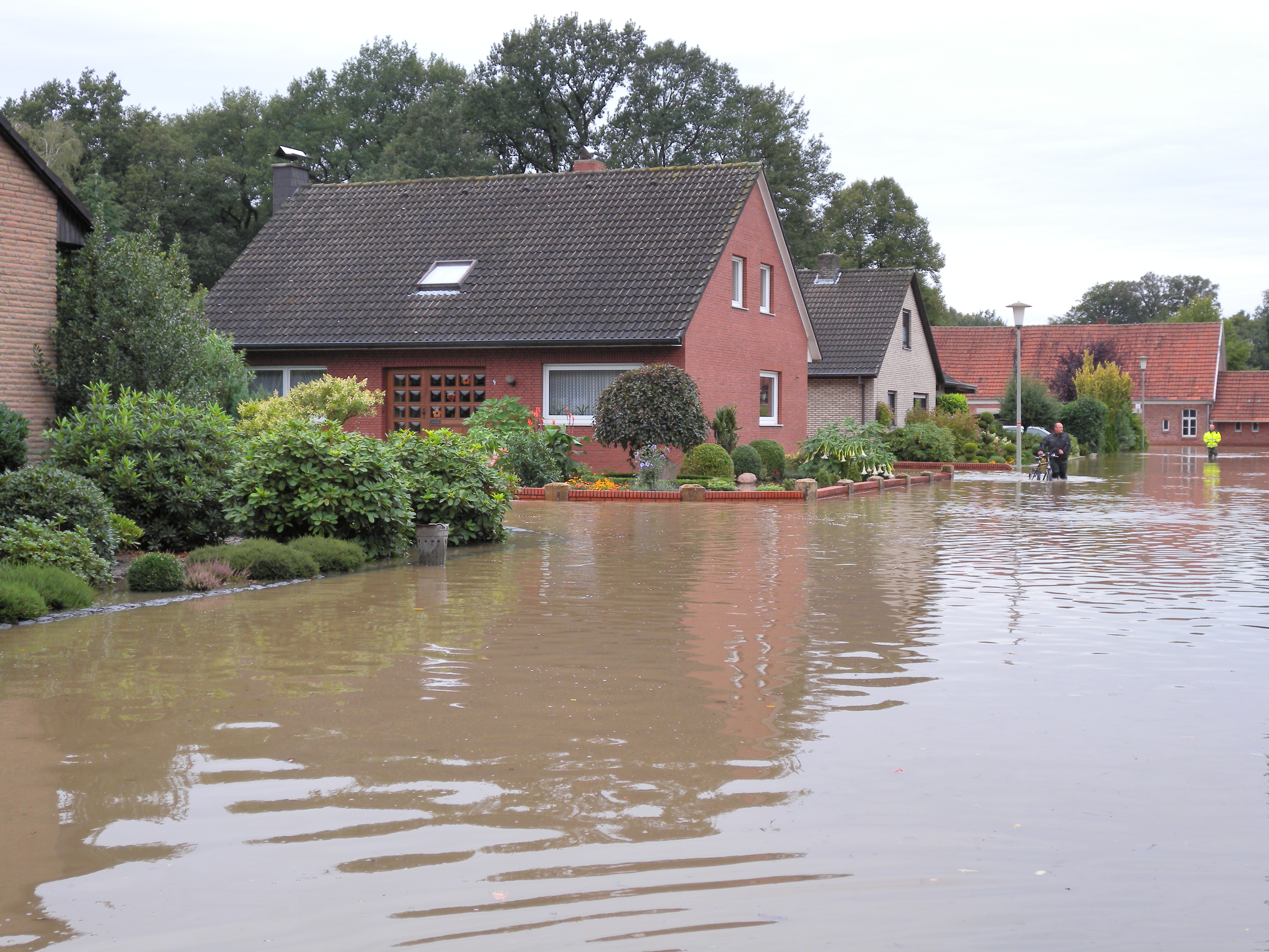 Auf dem Foto ist ein Wohngebiet mit freistehenden Einfamilienhäusern zu sehen. Die gesamte Straße, die etwas tiefer als die Grundstücke liegt, ist überschwemmt. Auch in den Vorgärten steht bereits das Wasser. Im Hintergrund waten zwei Menschen durch das kniehohe Wasser.