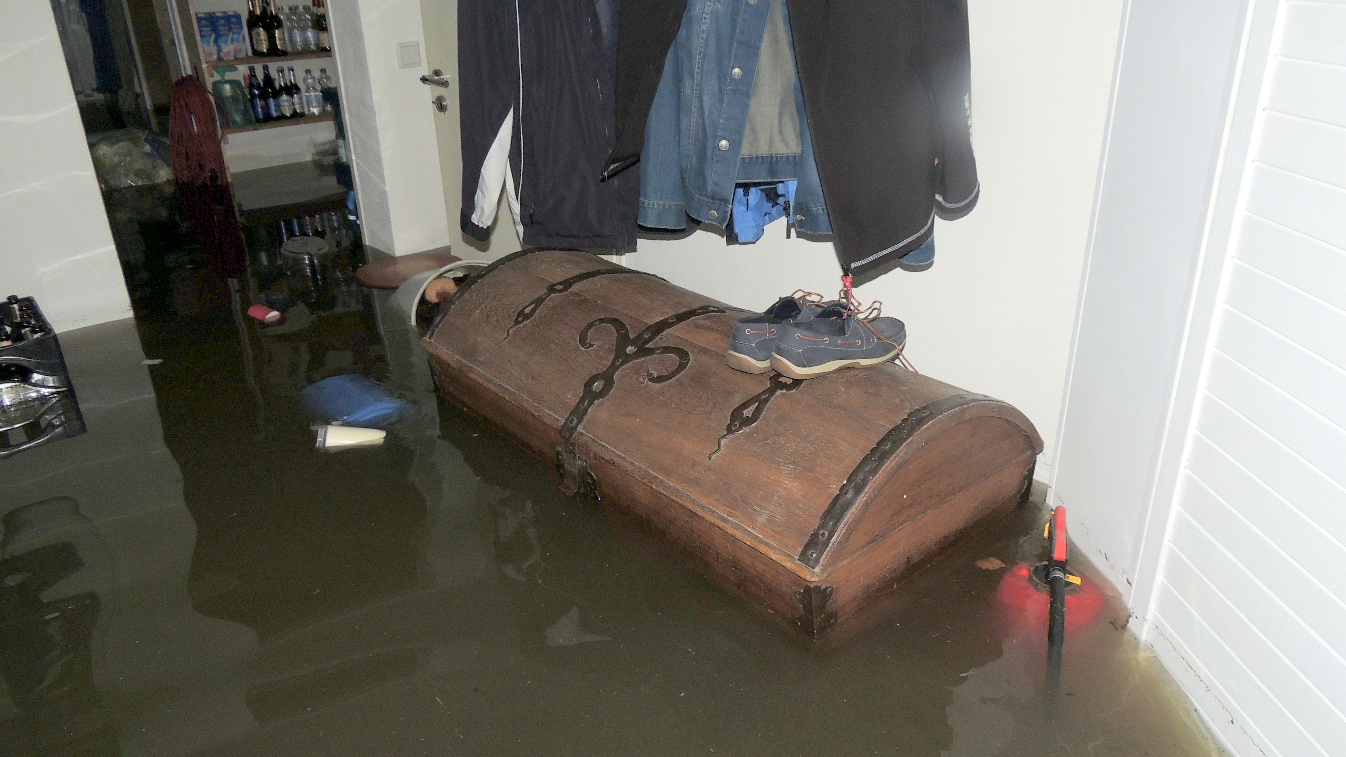 Das Foto zeigt einen Kellerraum einer Wohnung mit Garderobe, an der mehrere Jacken hängen. Darunter stehen eine große Holztruhe sowie ein Feuerlöscher. Die Truhe sowie der Feuerlöscher stehen fast vollständig unter Wasser. Weiterhin schwimmen vereinzelte Gegenstände, wie z.B. Schuhe im etwa einem Meter hohen Wasser.