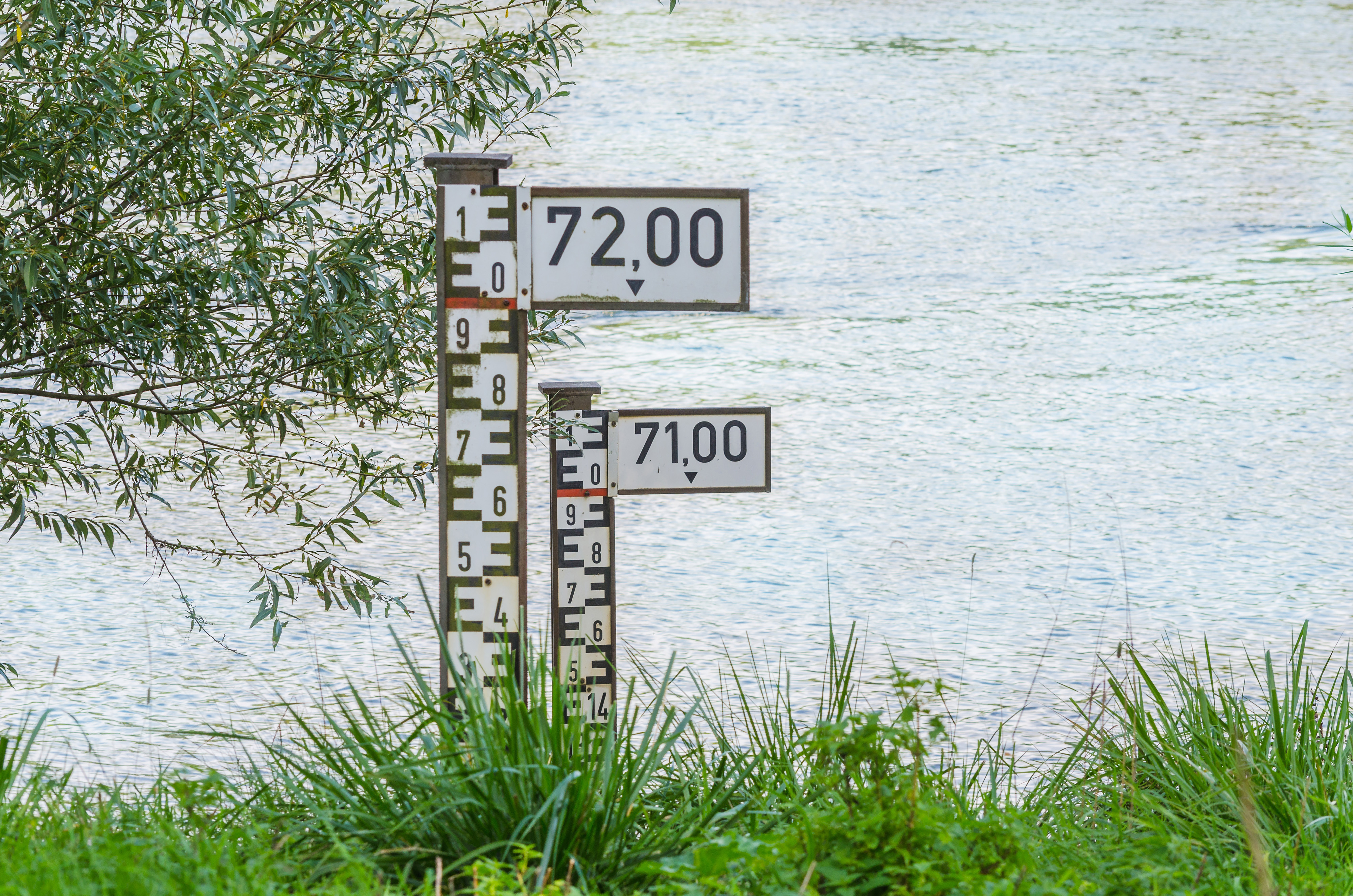 Das Foto zeigt eine Uferböschung mit dahinterliegendem Gewässer. Im Vordergrund befinden sich zwei Pegel, d.h. fest installierte Messlatten zur Feststellung des aktuellen Wasserstandes in Gewässern. Der vordere Pegel zeigt eine Höchstmarke von 72, was für eine Höhe von 7,20 m steht. Der hintere, sich näher am Gewässer befindende Pegel, hat eine Höchstmarke von 71. Den derzeitigen Wasserstand des Gewässers kann man nicht ablesen, da diese von der Vegetation der Uferböschung verdeckt wird.