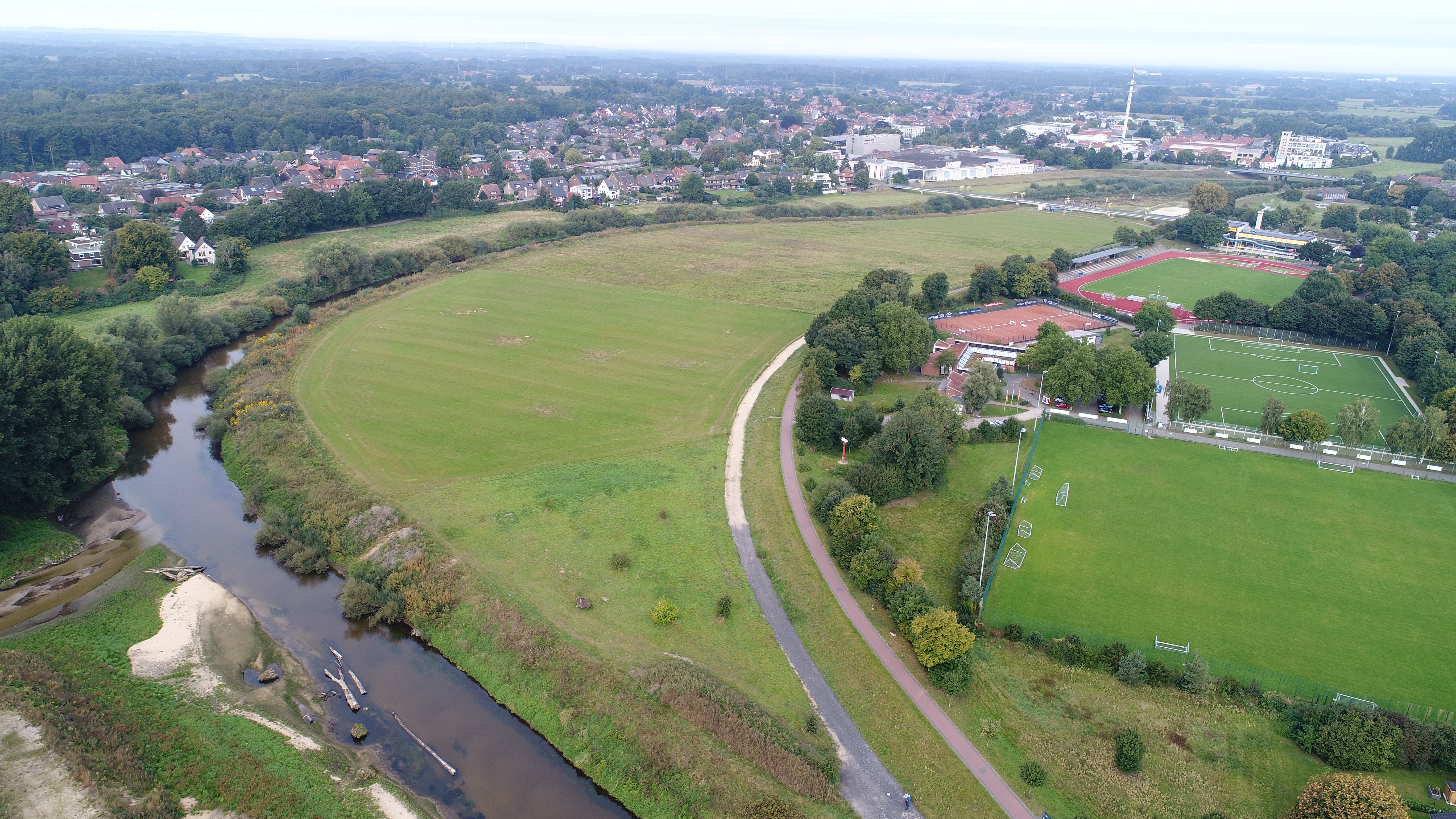 Luftbild mit Blick über die Baufläche vor Beginn der Arbeiten. Im Vordergrund links ist der umgestaltete Mündungsbereich der Münsterschen Aa zu sehen, im Hintergrund die Fußgängerbrücke am Hallenbad in Greven 