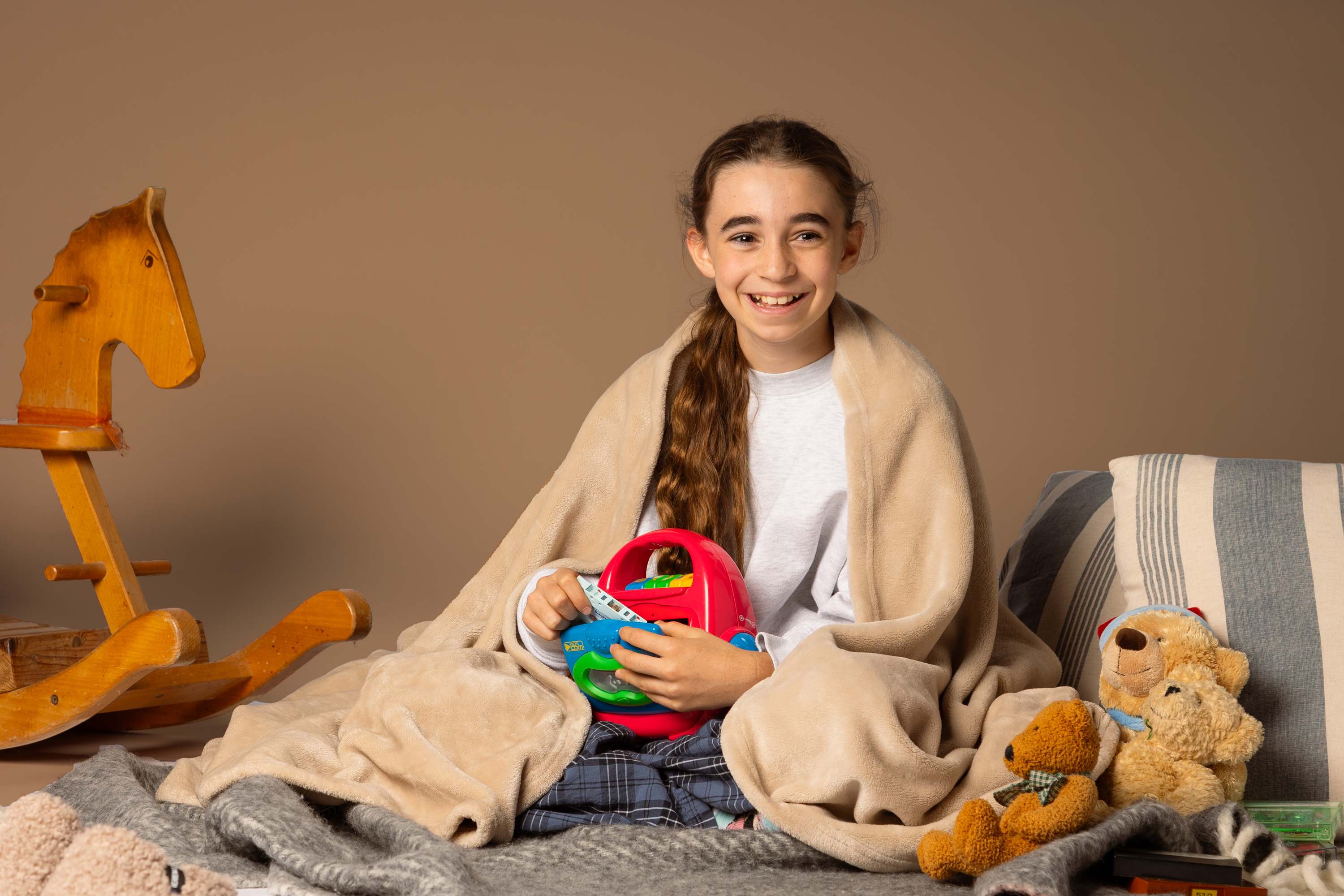 Ein Mädchen sitzt auf einer Decke und auch zugedeckt. Sie hält einen Kassettenrecorder. Neben ihr sind Teddybären