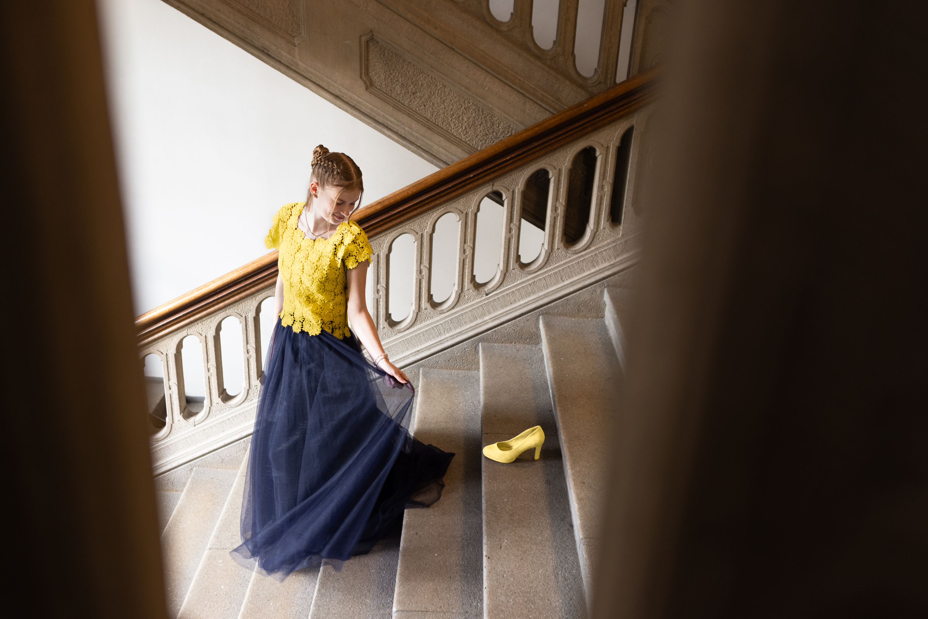 Eine weibliche Jugendliche geht in einem festlichen Kleid in Europafarben eine Treppe hinunter und schaut auf einen Schuh, der dort steht.