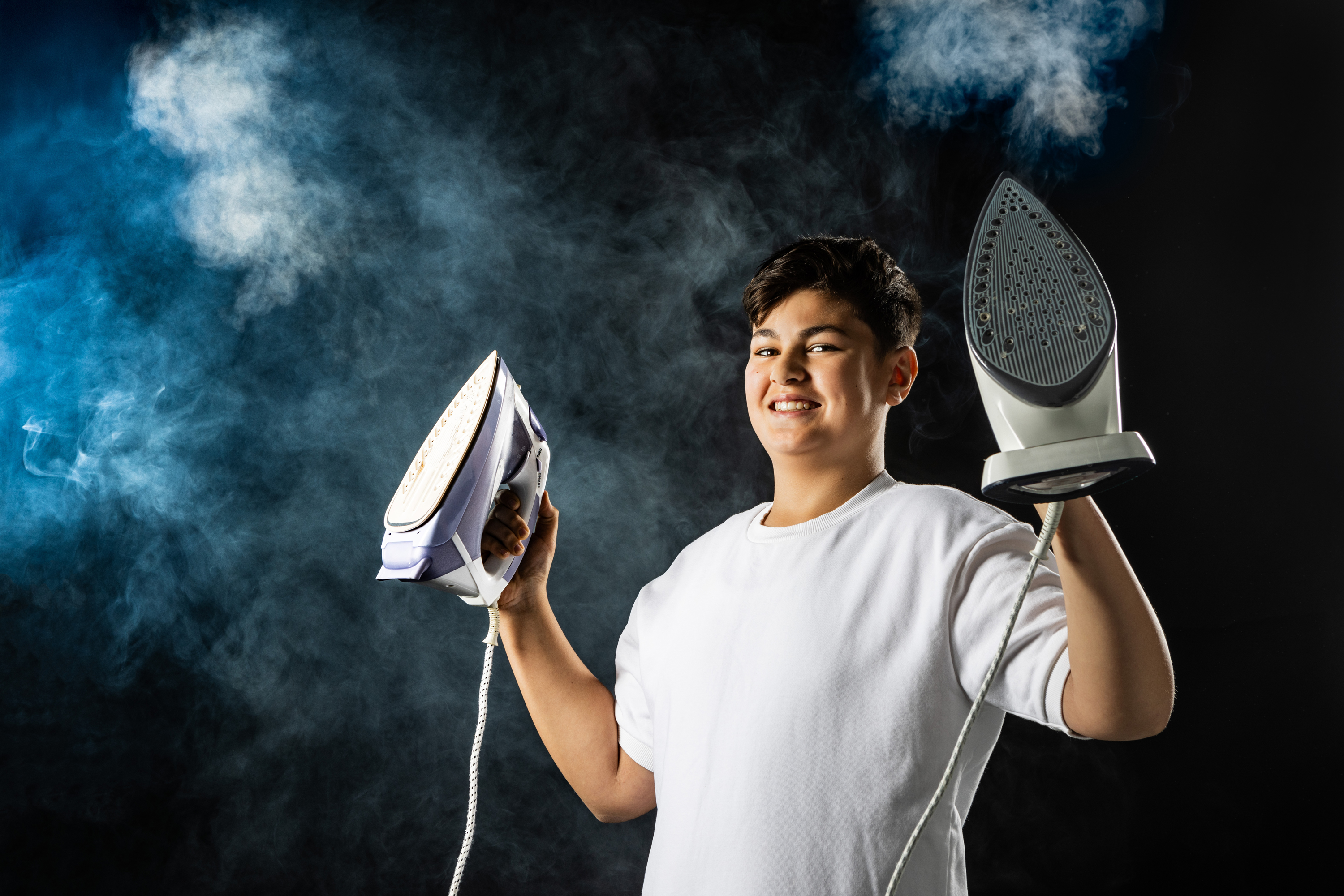 Ein männlicher Jugendlicher hält zwei Dampfbügeleisen hoch. Im Hintergrund ist Dampf und Nebel zu sehen.