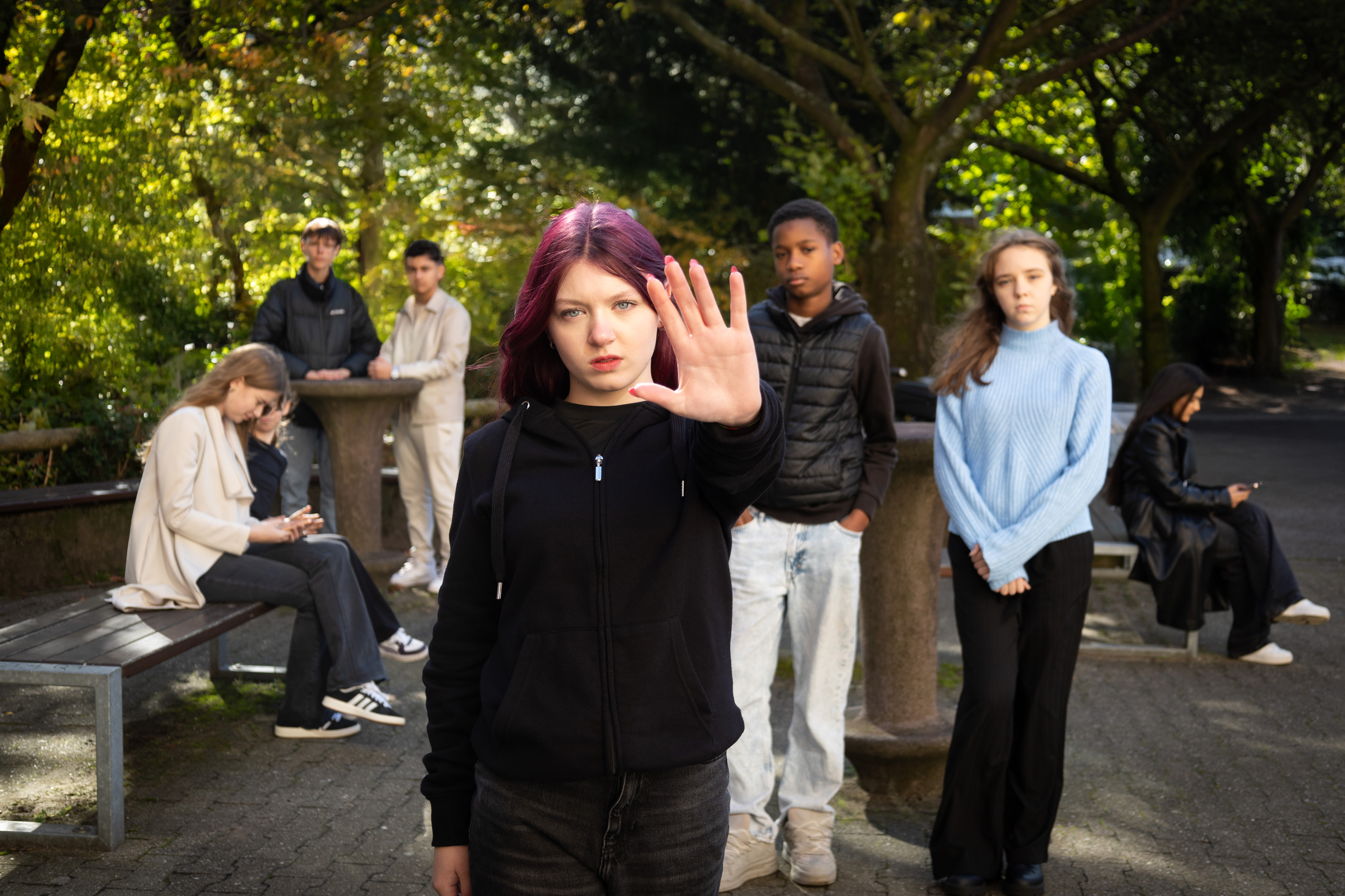 Sieben weibliche und männliche Jugendliche sind auf einem Schulhof. Die Jugendliche im Vordergrund macht eine Geste für "Stopp".