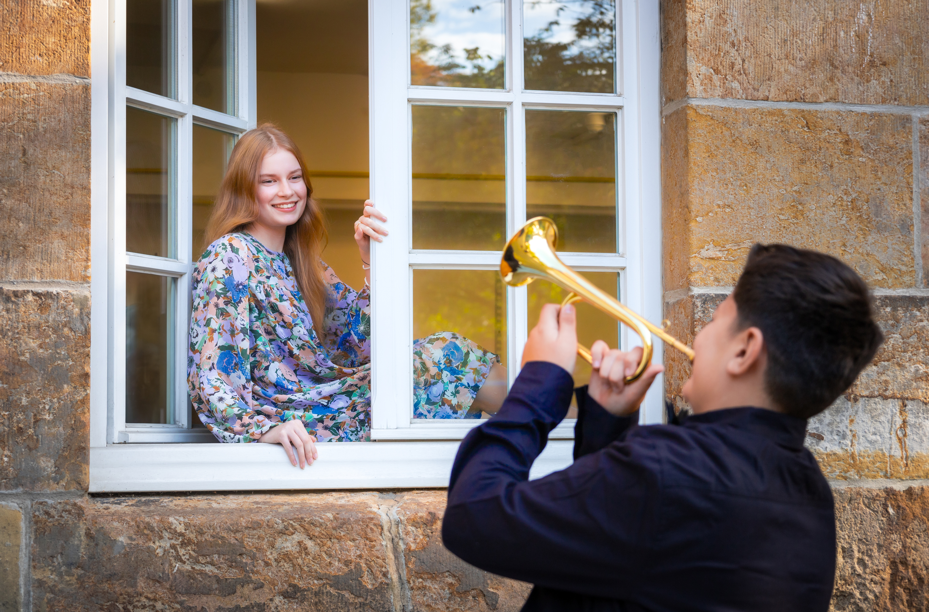Eine weibliche Jugendliche sitzt in einem Fenster, davor ein männlicher Jugendlicher der Trompete spielt.
