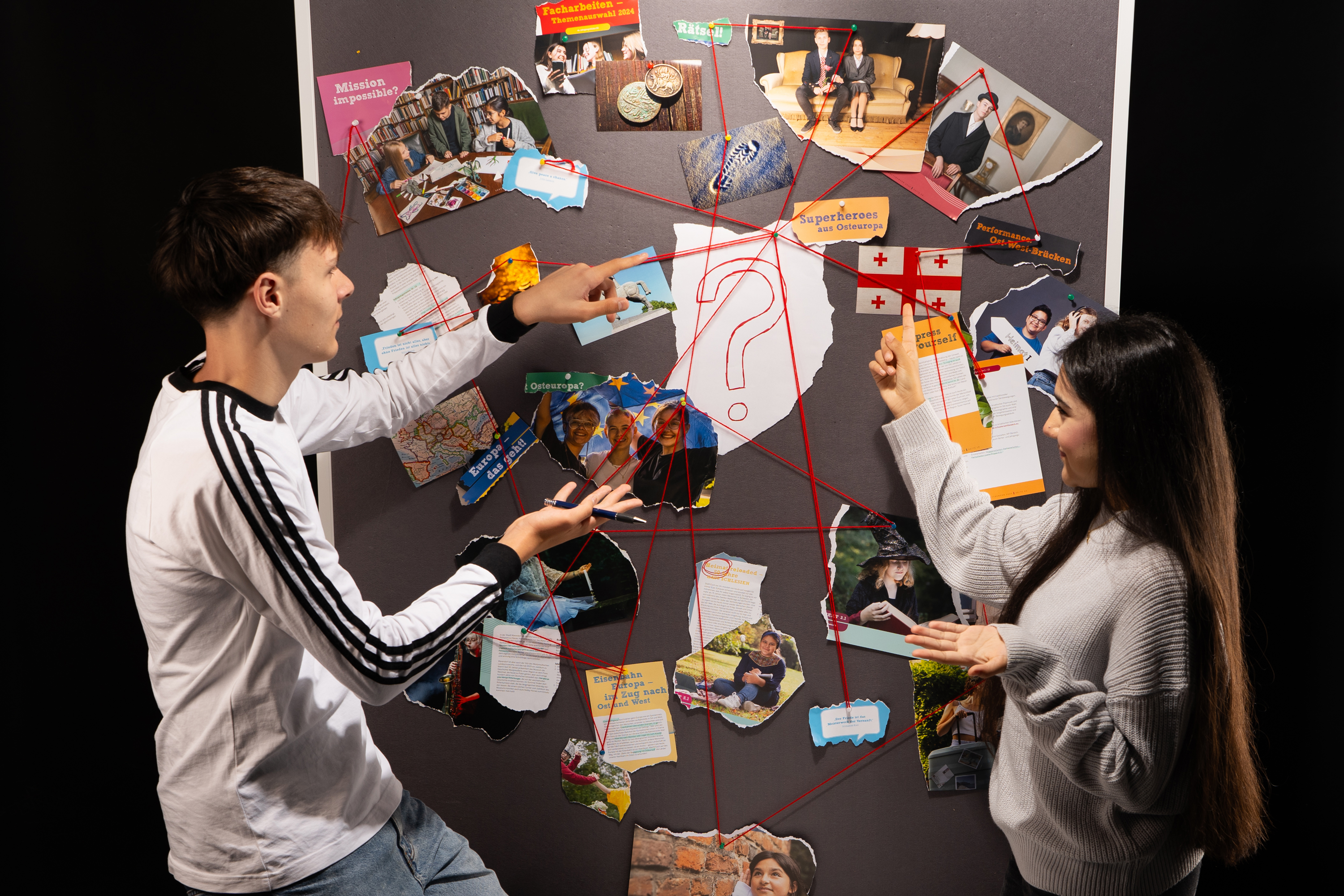 Zwei Jugendliche sehen vor einer Stellwand mit verschiedenen Fotos. Die Fotos sind mit roten Schnüren verbunden.