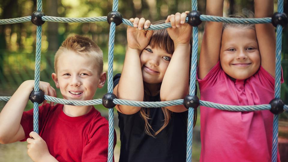 drei Kinder halten stehen hinter einem Spielgerät auf einem Spielplatz
