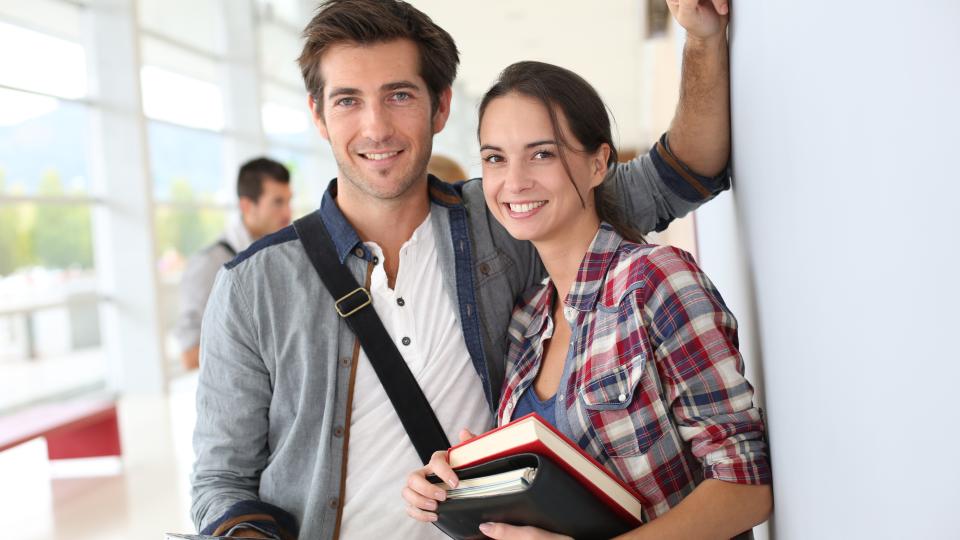 Ein junger Mann steht links, eine junge Frau steht rechts. Beide haben Bücher in den Händen und lächeln an der wand angelehnt in die Kamera.