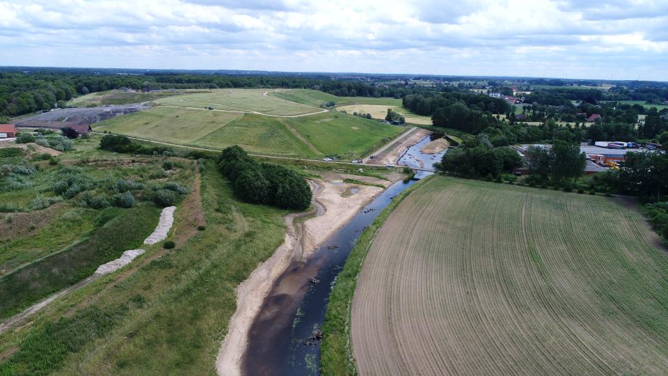 Zu sehen ist eine Drohnenaufnahme eines Abschnitts der Bocholter Aa in Bereich der Deponie Hoxfeld, die sich im rechten Bildhintergrund befindet. Der Gewässerverlauf wurde linksseitig aufgeweitet und Totholzelemente eingebaut, um die Bildung von naturnahen Habitatstrukturen zu fördern. 