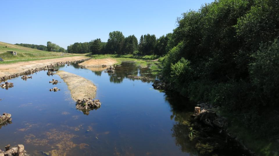 Zu sehen ist ein Abschnitt der Bocholter Aa in Hoxfeld, in dem strukturverbessernde Maßnahmen umgesetzt wurden. Mehrere Wurzelstubben wurden eingebaut und Inseln angelegt, um die Strukturvielfalt zu erhöhen. Links angrenzend an die Bocholter Aa ist die Deponie Hoxfeld zu erkennen, am rechten Ufer ist ein Gehölzsaum entlang der Aa zu sehen. 