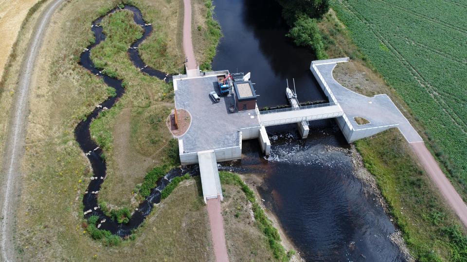 Zu sehen ist eine Drohnenaufnahme der Wehranlage Eisenhütte in der Bocholter Aa. Im zentralen Bildbereich befindet sich der Turbinenkanal der Wasserkraftanlage am rechten Ufer. Unmittelbar unterhalb des Turbinenkanals ist der Einstieg in die Fischaufstiegsanlage zu sehen, die in Riegelbauweise ausgeführt wurde.