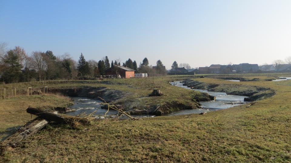 Zu sehen ist ein in Mäandern angelegtes Umgehungsgerinne zur Herstellung der Durchgängigkeit an der Plagemanns Mühle in Metelen in Riegelbauweise. Im Hintergrund ist die Wohnbebauung von Metelen zu sehen. 
