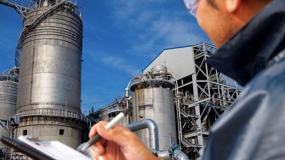 Eine Person mit einem Klemmbrett steht vor einer Industrieanlage