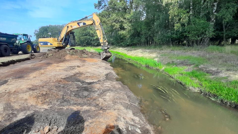 Ein Bagger nimmt Abgrabungen am Ufer eines Baches vor. Hinter dem Bagger befindet sich ein Traktor mit Anhänger, der die Erde abtransportiert. 