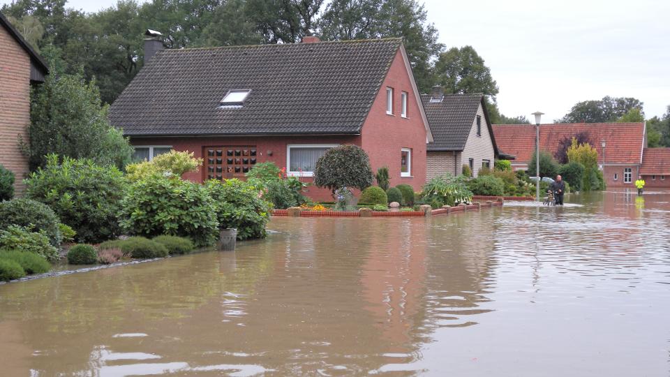 Auf dem Foto ist ein Wohngebiet mit freistehenden Einfamilienhäusern zu sehen. Die gesamte Straße, die etwas tiefer als die Grundstücke liegt, ist überschwemmt. Auch in den Vorgärten steht bereits das Wasser. Im Hintergrund waten zwei Menschen durch das kniehohe Wasser.