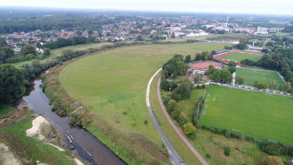 Luftbild mit Blick über die Baufläche vor Beginn der Arbeiten. Im Vordergrund links ist der umgestaltete Mündungsbereich der Münsterschen Aa zu sehen, im Hintergrund die Fußgängerbrücke am Hallenbad in Greven 
