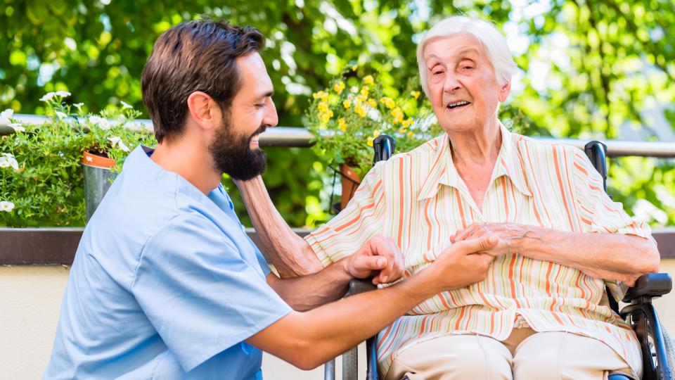 Ein männlicher Pfleger kniet neben dem Rollstuhl einer älteren Dame