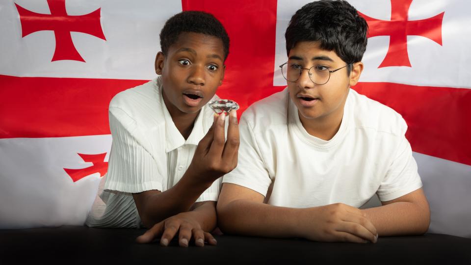 Zwei Jungen schauen auf einen Diamanten, den einer der Jungen in der Hand hält. Im Hintergrund ist die Fahne von Georgien