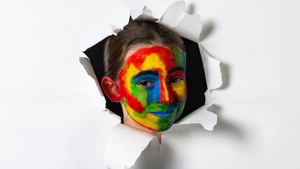 Ein Mädchen mit einem bunt bemalten Gesicht schaut durch ein Loch in einer Leinwand.