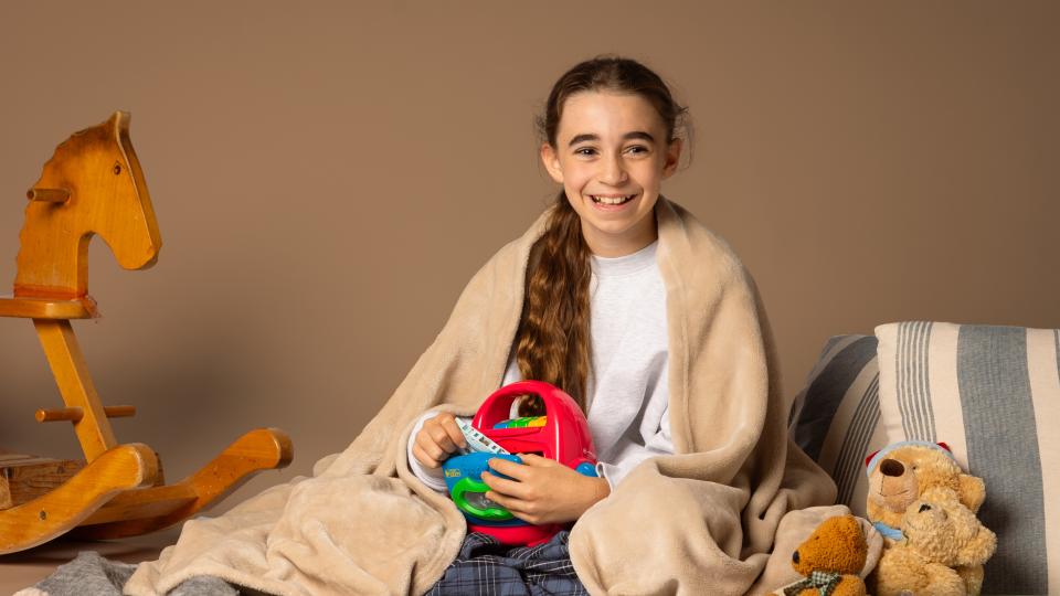 Ein Mädchen sitzt auf einer Decke und auch zugedeckt. Sie hält einen Kassettenrecorder. Neben ihr sind Teddybären