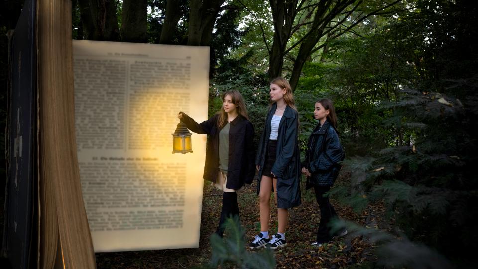 Fotocollage: Drei weibliche Jugendliche gehen auf ein hohes Buch zu. Eine hält eine Laterne in der Hand.