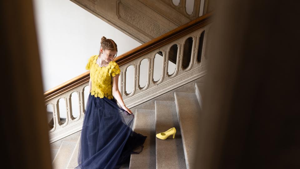 Eine weibliche Jugendliche geht in einem festlichen Kleid in Europafarben eine Treppe hinunter und schaut auf einen Schuh, der dort steht.