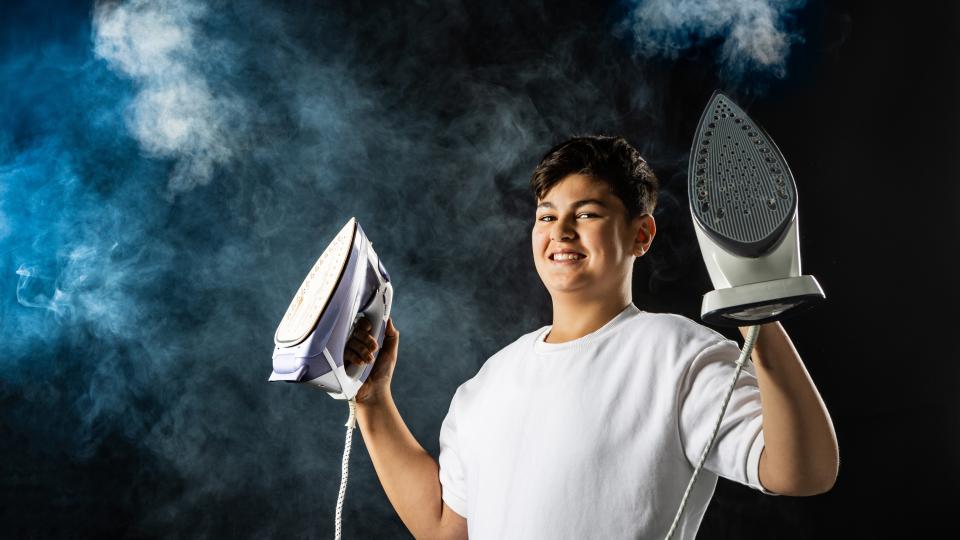 Ein männlicher Jugendlicher hält zwei Dampfbügeleisen hoch. Im Hintergrund ist Dampf und Nebel zu sehen.