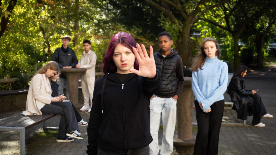 Sieben weibliche und männliche Jugendliche sind auf einem Schulhof. Die Jugendliche im Vordergrund macht eine Geste für "Stopp".