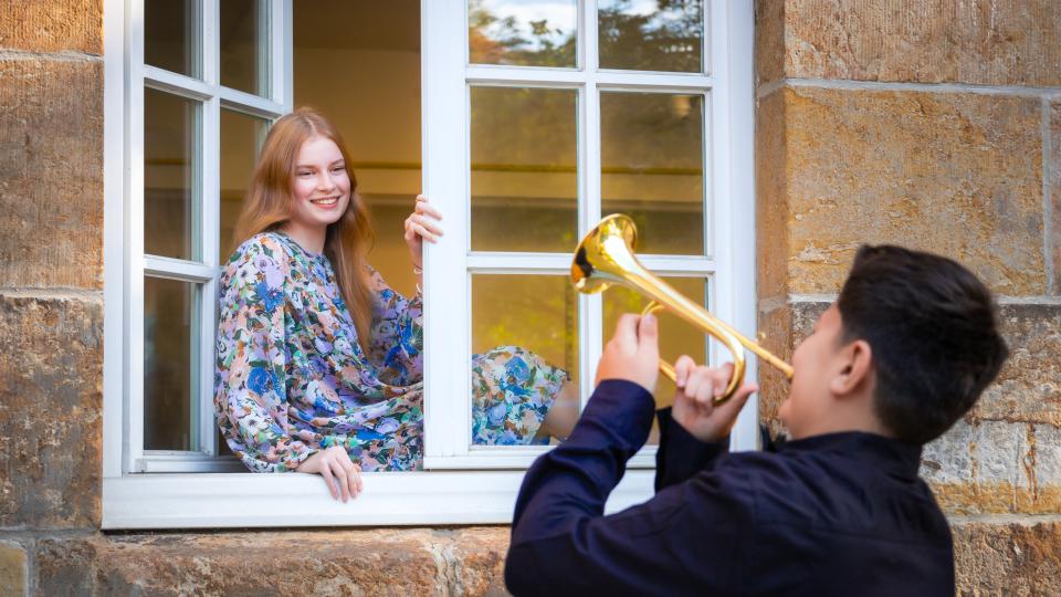 Eine weibliche Jugendliche sitzt in einem Fenster, davor ein männlicher Jugendlicher der Trompete spielt.