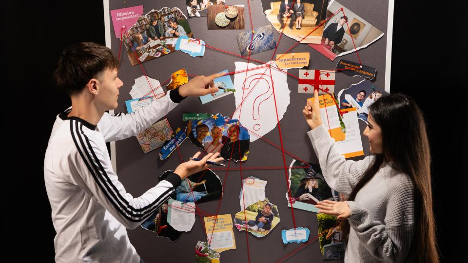 Zwei Jugendliche sehen vor einer Stellwand mit verschiedenen Fotos. Die Fotos sind mit roten Schnüren verbunden.