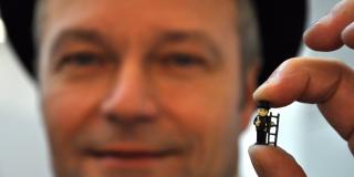 Gesicht eines Schornsteinfegers im Hintergrund unscharf, im Vordergrund hält er einen kleinen Plastikschornsteinfeger in die Kamera