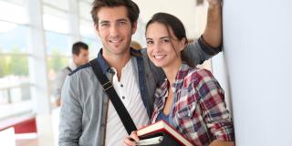 Ein junger Mann steht links, eine junge Frau steht rechts. Beide haben Bücher in den Händen und lächeln an der wand angelehnt in die Kamera.