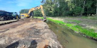 Ein Bagger nimmt Abgrabungen am Ufer eines Baches vor. Hinter dem Bagger befindet sich ein Traktor mit Anhänger, der die Erde abtransportiert. 