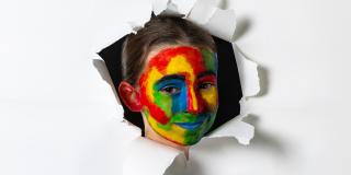 Ein Mädchen mit einem bunt bemalten Gesicht schaut durch ein Loch in einer Leinwand.
