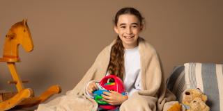 Ein Mädchen sitzt auf einer Decke und auch zugedeckt. Sie hält einen Kassettenrecorder. Neben ihr sind Teddybären