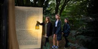 Fotocollage: Drei weibliche Jugendliche gehen auf ein hohes Buch zu. Eine hält eine Laterne in der Hand.