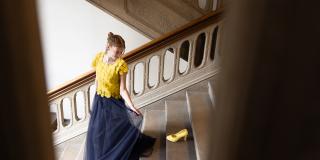 Eine weibliche Jugendliche geht in einem festlichen Kleid in Europafarben eine Treppe hinunter und schaut auf einen Schuh, der dort steht.