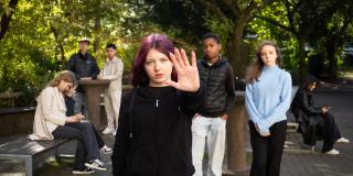 Sieben weibliche und männliche Jugendliche sind auf einem Schulhof. Die Jugendliche im Vordergrund macht eine Geste für "Stopp".