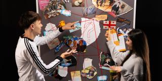 Zwei Jugendliche sehen vor einer Stellwand mit verschiedenen Fotos. Die Fotos sind mit roten Schnüren verbunden.